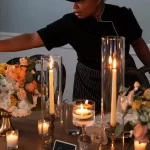 a woman in a black shirt and black hat standing at a table with flowers and candles setting up a luxury event